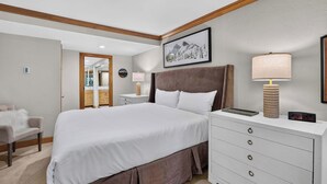 A hotel room with a large bed, a nightstand with a lamp, a dresser, and a framed picture on the wall. - Lodge at Lionshead 210A (Vail)