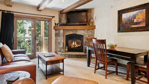 A cozy living room with a fireplace, a television mounted above it, and a dining area with chairs and a table. - Lodge at Lionshead 230B (Vail)