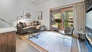 Un salon moderne avec un canapé, une table basse et un fauteuil. Il y a une grande fenêtre avec des rideaux, un ventilateur de plafond et un téléviseur à écran plat.