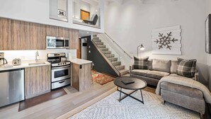 Cuisine moderne équipée d’un four à micro-ondes, d’un grille-pain et d’un four traditionnel. Un escalier mène à l’étage supérieur. Le salon comprend un canapé et une table basse.