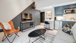 A modern living room with a sofa, a coffee table, and a TV. There is a kitchen area with cabinets and a microwave. - Lodge at Lionshead 330 (Vail)