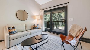 A modern living room with a sofa, a coffee table, a chair, a round mirror, and a sliding glass door leading to an outdoor area. - Lodge at Lionshead 330 (Vail)