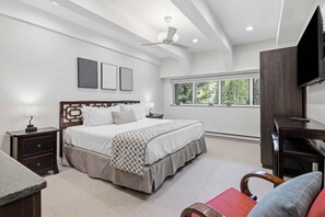 A bedroom with a bed, bedside tables, a desk, a chair, a TV, and a window with a view of trees. - Lodge at Lionshead 350 (Vail)