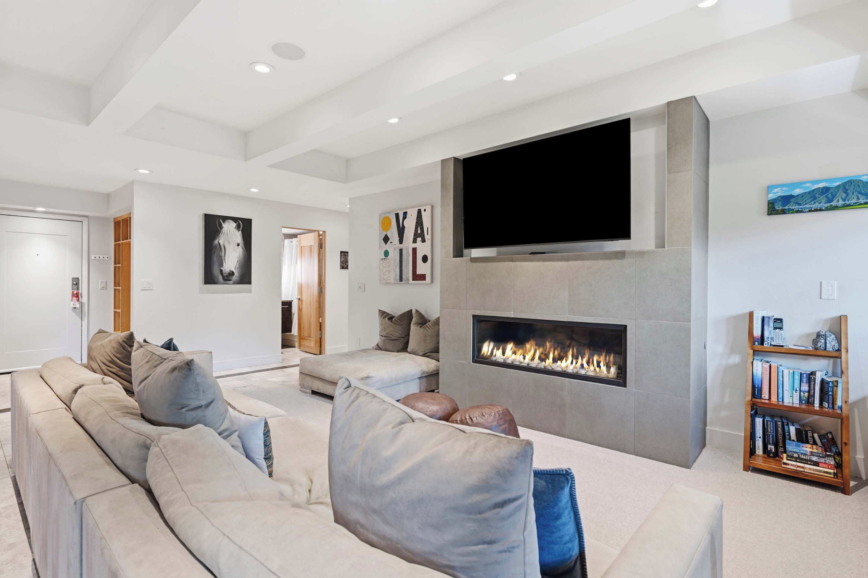 A modern living room with a large sectional sofa, a flat-screen TV mounted on a stone wall, a built-in fireplace, and a bookshelf.