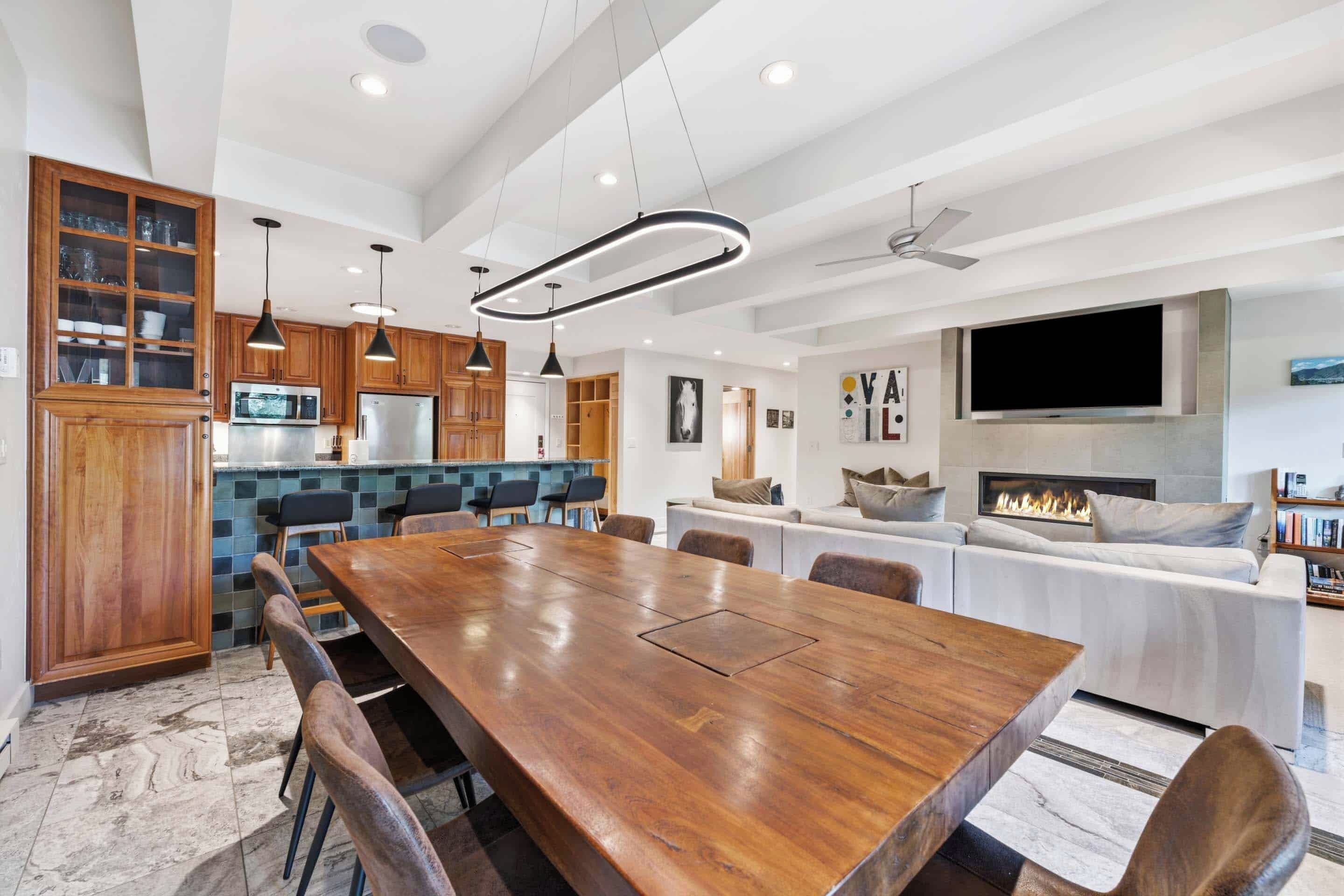A modern kitchen with wooden cabinets, a dining area with a large wooden table, and a living room with a fireplace and a flat-screen TV.