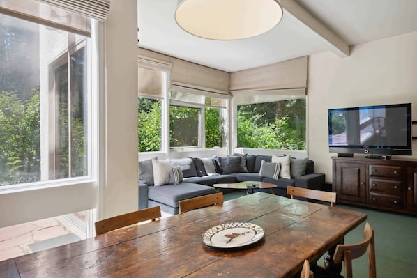 A modern living room with a large window, a sofa, a dining table, and a television.