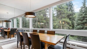 Un comedor moderno con una mesa grande y sillas, ventanas amplias que dan a vista de árboles, e iluminación colgante.
