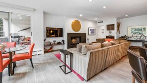 Una sala de estar moderna con un sofá modular, una mesa de comedor de vidrio con sillas anaranjadas, una chimenea y un ventanal grande.