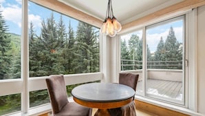 A round wooden table with two upholstered chairs in a room with large windows overlooking a forest.