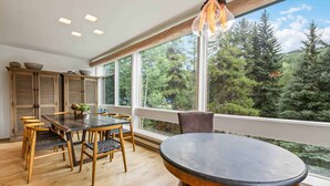 A dining area with a round table and chairs, wooden cabinets, and large windows offering a view of trees.