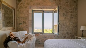 A room with a stone wall, a large window, a bed, and a chair. - Riglia Villas - Villa Olive (West Mani)