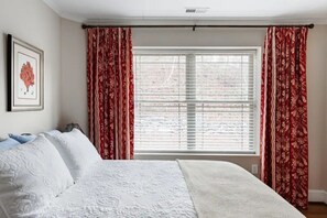 Ein Schlafzimmer mit einem großen Bett, weißer Bettwäsche, einem gerahmten Bild an der Wand und roten, gemusterten Vorhängen.