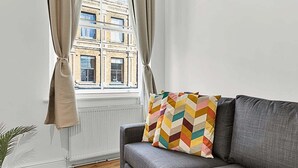 A modern living room with a grey sofa, a colorful chevron-patterned pillow, a potted plant, and a view of a brick building through a window with beige curtains.