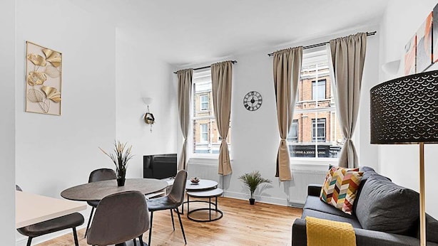 A modern living room with a dining area, large windows with curtains, a clock, and a sofa with a colorful pillow.