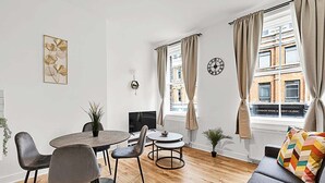A modern living room with a round dining table, two chairs, a sofa, a potted plant, and a view of a building with a sign reading "LONDON SPINE & JOINT CLINIC".