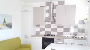 A modern kitchen with white cabinets, a grey and white checkered backsplash, a stainless steel range hood, and a dining area with a white table and metal chairs. - Holiday home with air conditioning and washing machine (Rosolina Mare)