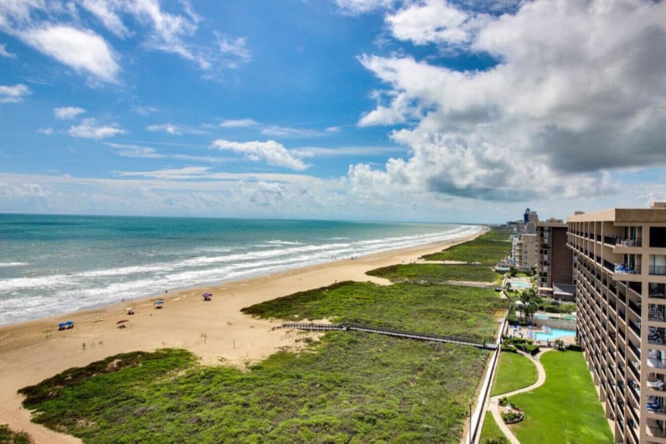 A beachfront hotel with a view of the ocean, a sandy beach, and a grassy area with a walking path.