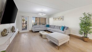 Una sala moderna con un sofá seccional gris, un televisor de pantalla plana montado en la pared, una planta grande en maceta y un balcón con vistas a la ciudad.