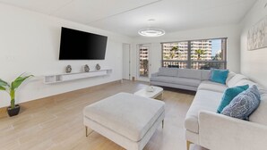 Una sala de estar moderna con un televisor de pantalla plana, un sofá gris, un otoman blanco y una planta en maceta.