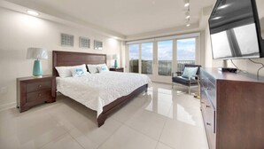 A modern bedroom with a large bed, a TV, and a balcony with a city view. - Luxury Beachfront Condo With Amazing Sunset Views (South Padre Island)