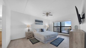 A modern bedroom with a bed, nightstands, a ceiling fan, a large window, and a view of the city.
