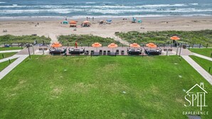 Une vue sur la plage, avec une zone herbeuse, des espaces de détente en plein air dotés de parasols et un ciel dégagé.