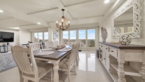 A dining area with a large wooden table, white chairs, a chandelier, and a view of the city through large windows.