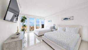Ein modernes Schlafzimmer mit zwei Betten, einem großen Fenster mit Blick auf den Strand und einem an der Wand montierten Flachbildfernseher.