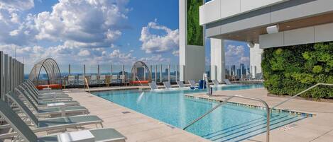 A rooftop pool with lounge chairs, a view of the city skyline, and a green wall.