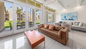 A spacious living room with a large window, a brown leather sofa, a wooden coffee table, and a grey sofa.