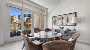 A modern dining area with a glass table surrounded by wicker chairs, large windows offering a view of a pool and buildings, and a contemporary painting on the wall.