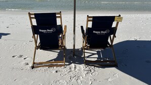 Två strandstolar under ett stort parasoll på en sandstrand, med fotspår som leder ner mot vattnet.