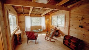 Interior de una cabaña de madera con una mecedora, una mesita y un cofre rojo.