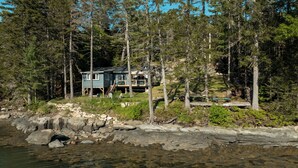 Eine Hütte, umgeben von dichtem Wald, mit felsiger Küste und klarem Wasser im Vordergrund.