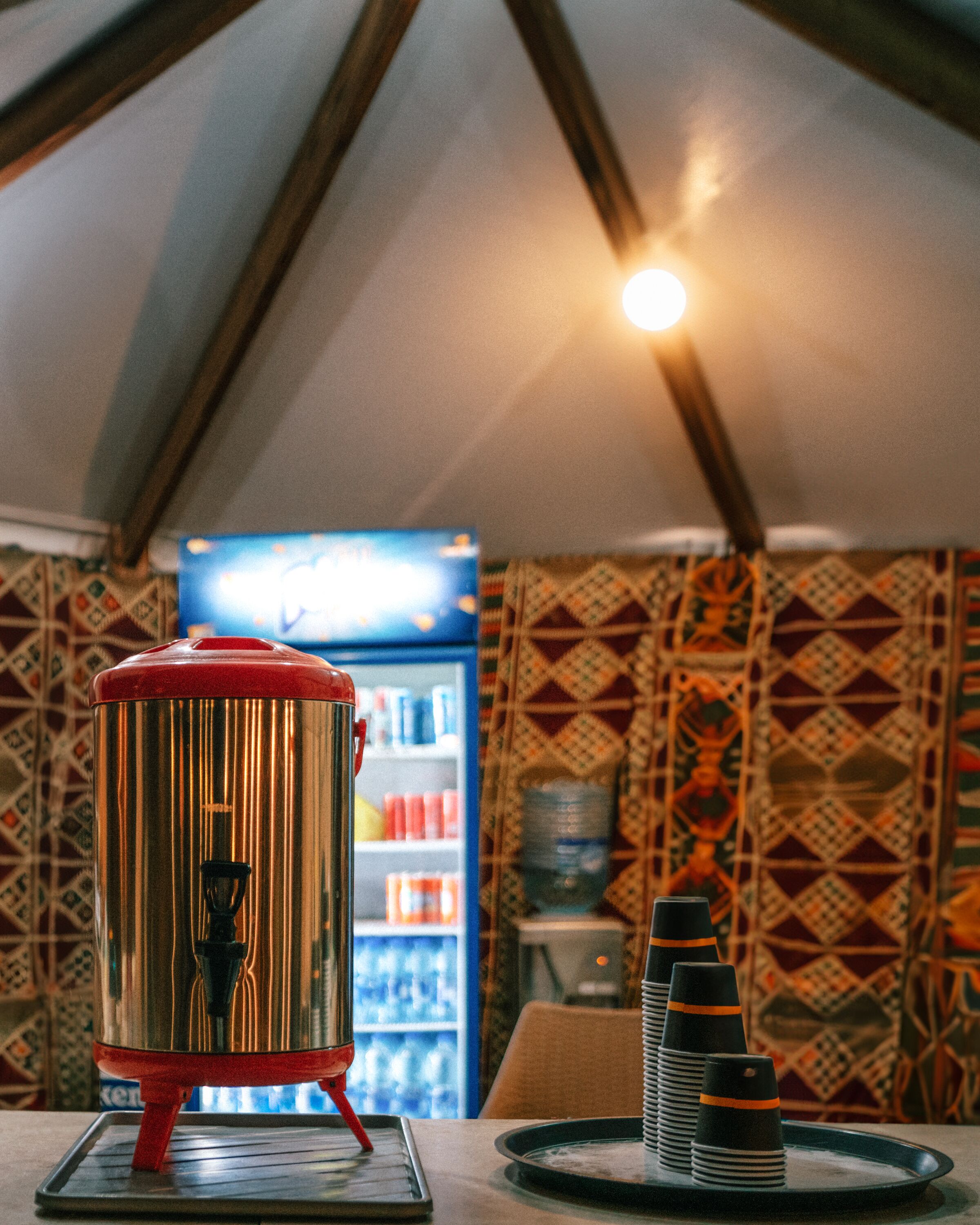 A kitchen area with a stainless steel coffee maker, a refrigerator stocked with beverages, and a patterned wall.