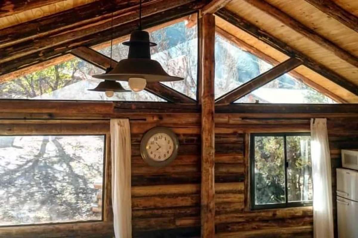 A rustic cabin interior with a wooden ceiling, a hanging light fixture, a clock, and a refrigerator.