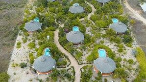 Luchtbeeld van een resort met hutten met rieten daken, gerangschikt in een halve cirkel rond een hoofdpad, dat naar een strand leidt.