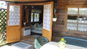 En træhytte med veranda, et bord med en vase blomster, og et vindue med blonde gardiner.