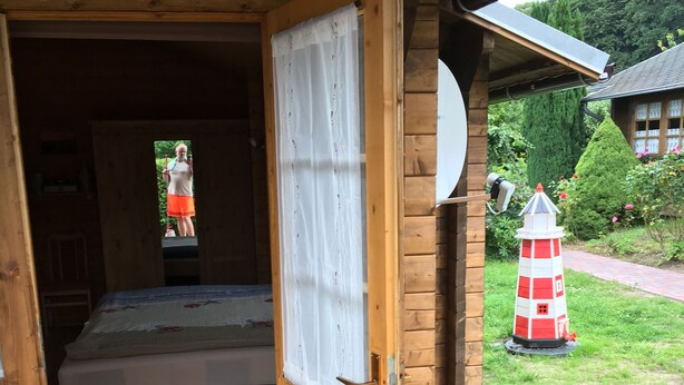 Une cabane en bois avec un lit, une petite véranda et un jardin agrémenté de fleurs et d’une maquette de phare.