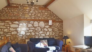 A cozy living room with a stone wall, wooden beams, a chandelier, a sofa set, a side table with a lamp, and a TV stand with books. - 2 bedroom accommodation in Oswestry (Oswestry)