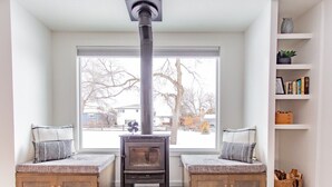 A cozy room with a wood-burning stove, a bench with cushions, and a window with a view of trees and a house. - Bridger Hawk Duplex (NEW) (Bozeman)