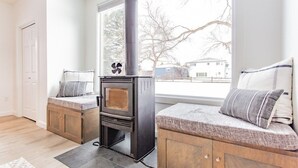 A cozy room with a wood-burning stove, a bench with cushions, and a view of a tree outside. - Bridger Hawk Duplex (NEW) (Bozeman)
