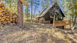 A rustic cabin with a wooden deck, surrounded by trees and a stack of firewood. - Ruidoso Rental w/ Private Hot Tub: Walk to Midtown (Ruidoso)