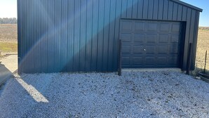 Metallinen varastorakennus, jossa on autotallin ovi, sorapäällysteinen ajotie ja kirkas taivas.