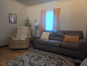 Uma sala de estar com um sofá de couro marrom, uma poltrona bege, um tapete estampado, um vaso de planta e um quadro pendurado na parede.