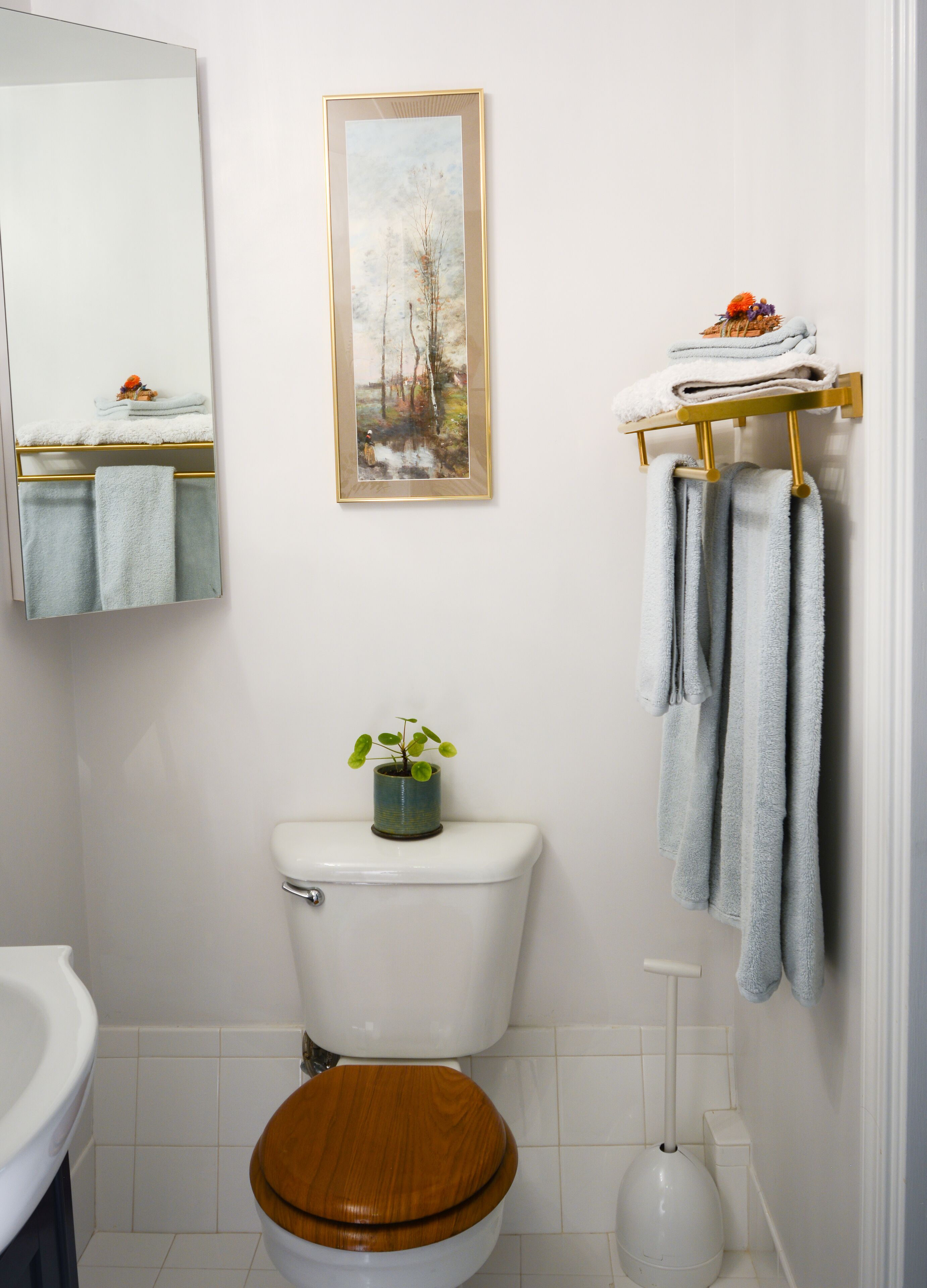 A bathroom with a wooden toilet, a white sink, a framed painting, a towel rack with towels, and a small potted plant.