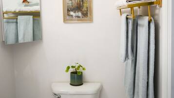 A bathroom with a wooden toilet, a white sink, a framed painting, a towel rack with towels, and a small potted plant.