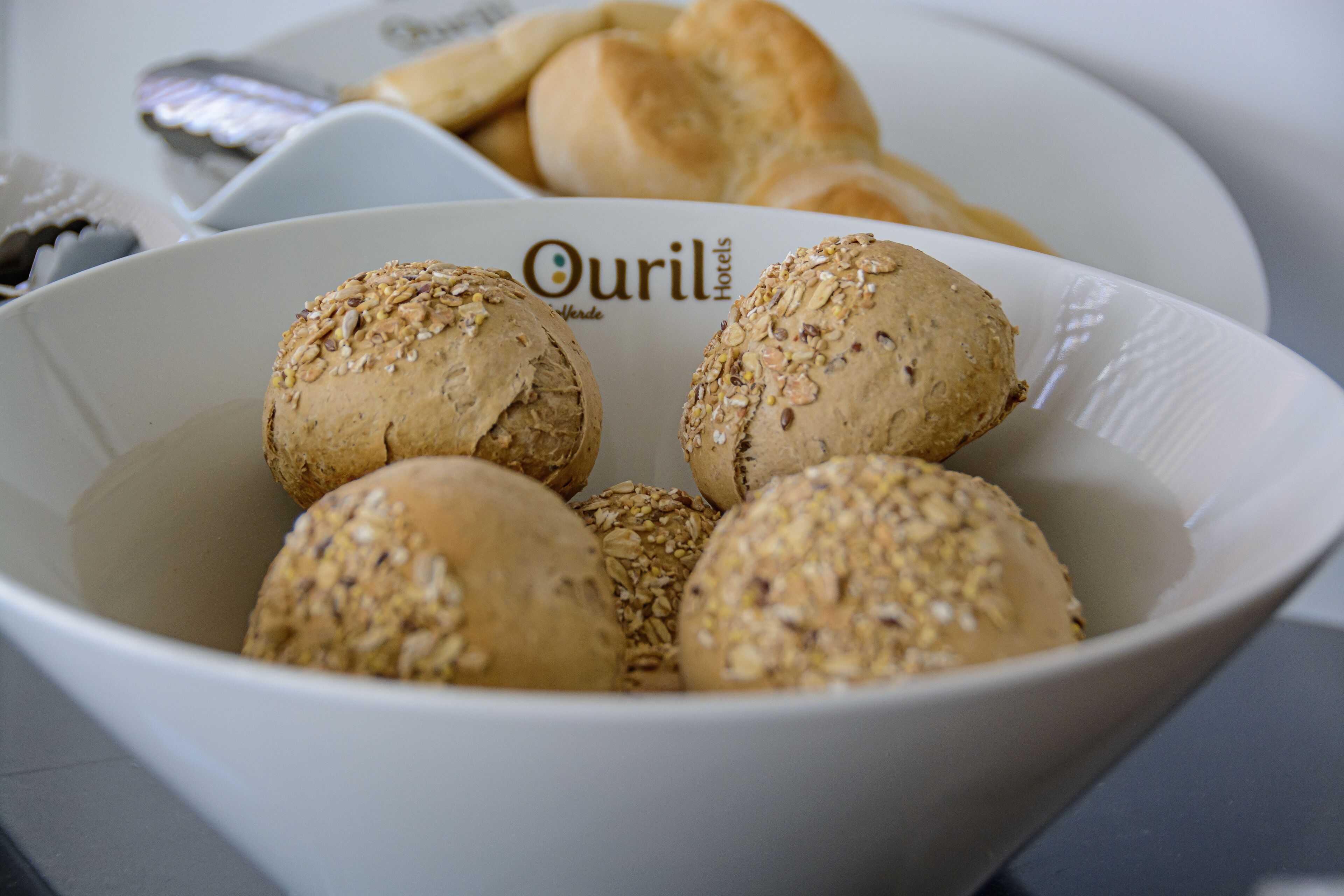 A bowl of assorted bread rolls with seeds, placed on a table with a white plate and a box labeled "Ouril Hotels" in the background.