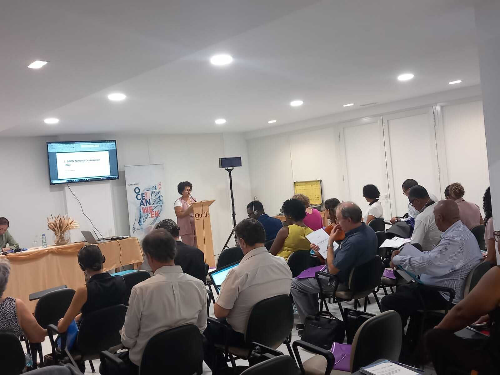 Une salle de conférence avec un conférencier à un pupitre, des participants assis à des tables et un écran de présentation.
