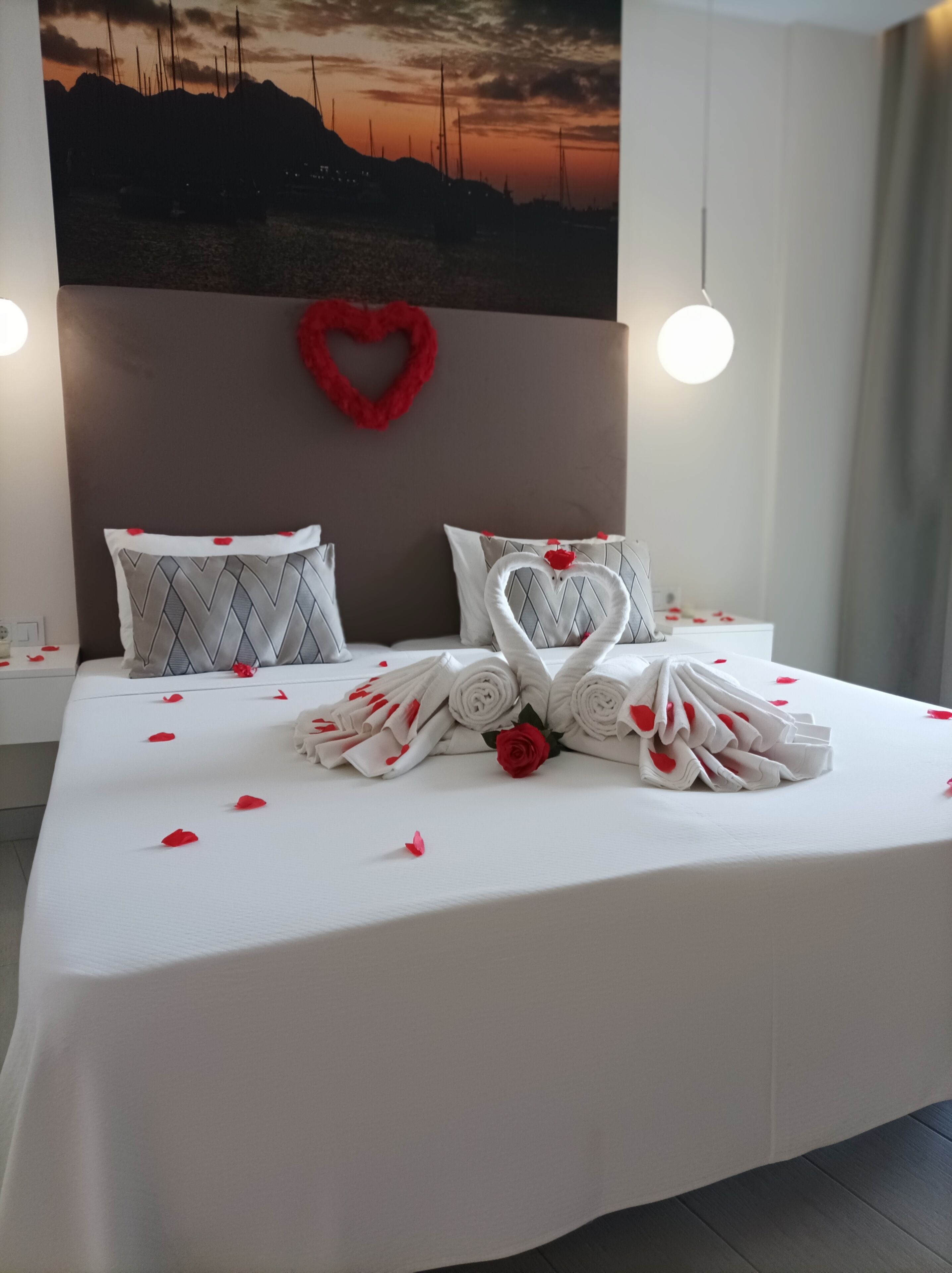 Un lit bien fait, avec des serviettes pliées et un coussin en forme de cœur, une décoration en forme de cœur rouge sur le mur et une vue sur le coucher de soleil.
